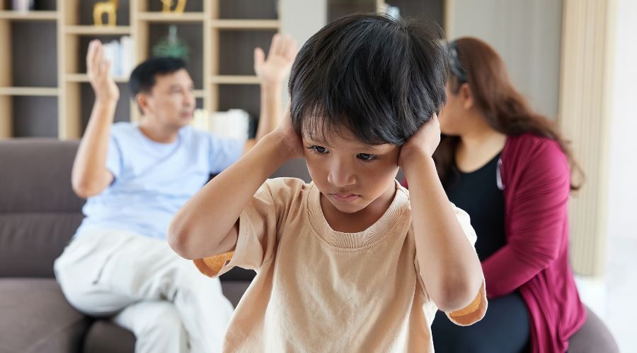 Child holding hands over ears with parents arguing behind him