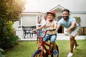 Father is teaching his child to ride a bike, and mother and daughter are watching nearby.