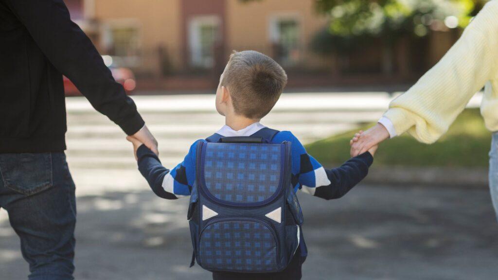 A young child wearing a blue backpack walks while holding hands with two adults, one on each side, on a sunny street.