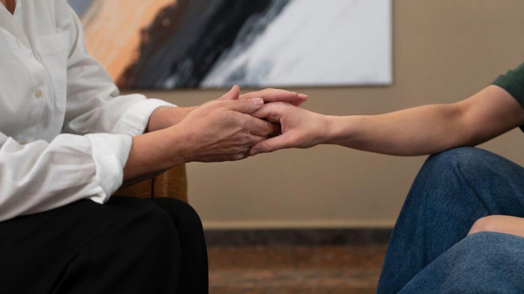 “Close-up of two people holding hands in a comforting gesture, one wearing a white shirt and the other in jeans, symbolizing support and compassion"