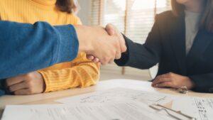 Two people shaking hands across a desk with legal documents and a clipboard, symbolizing agreement or partnership.