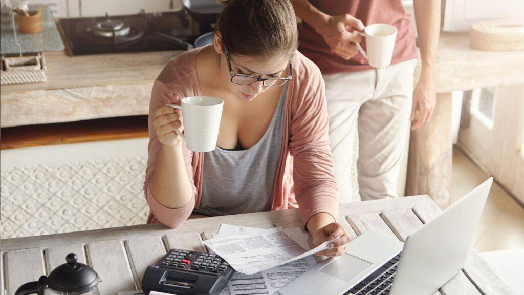 A woman sitting at a kitchen table holding a mug while reviewing documents, with a laptop and calculator in front of her, and another person standing nearby holding a mug.