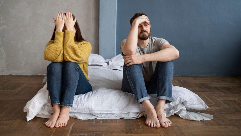 Unhappy couple sitting on the floor back-to-back near a bed, both looking stressed and upset, symbolizing relationship conflict.